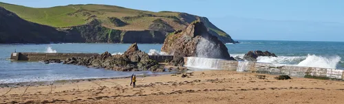 This landscape photograph depicts the beach wall at Hope Cove in Devon, England, where a wave is dramatically crashing against the coastal barrier. Taken in the morning during the spring season, the image shows the rugged coastline characteristic of the United Kingdom, with rolling green hills rising behind the bay and prominent rocky formations partially surrounded by the sea. Two people are visible walking across the sandy beach in the foreground, adding a sense of scale to the scene. The photograph captures the interaction between people and nature at this popular location on the English coast.
